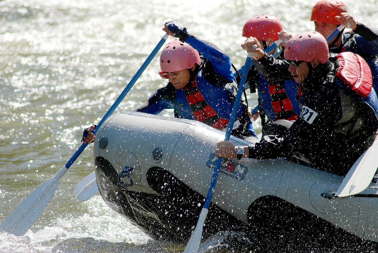 Eine Gruppe von Personen, die Helme und Schwimmwesten tragen, paddeln kräftig in einem Wildwasserfloß und spritzen Wasser, während sie durch einen reißenden Fluss navigieren.